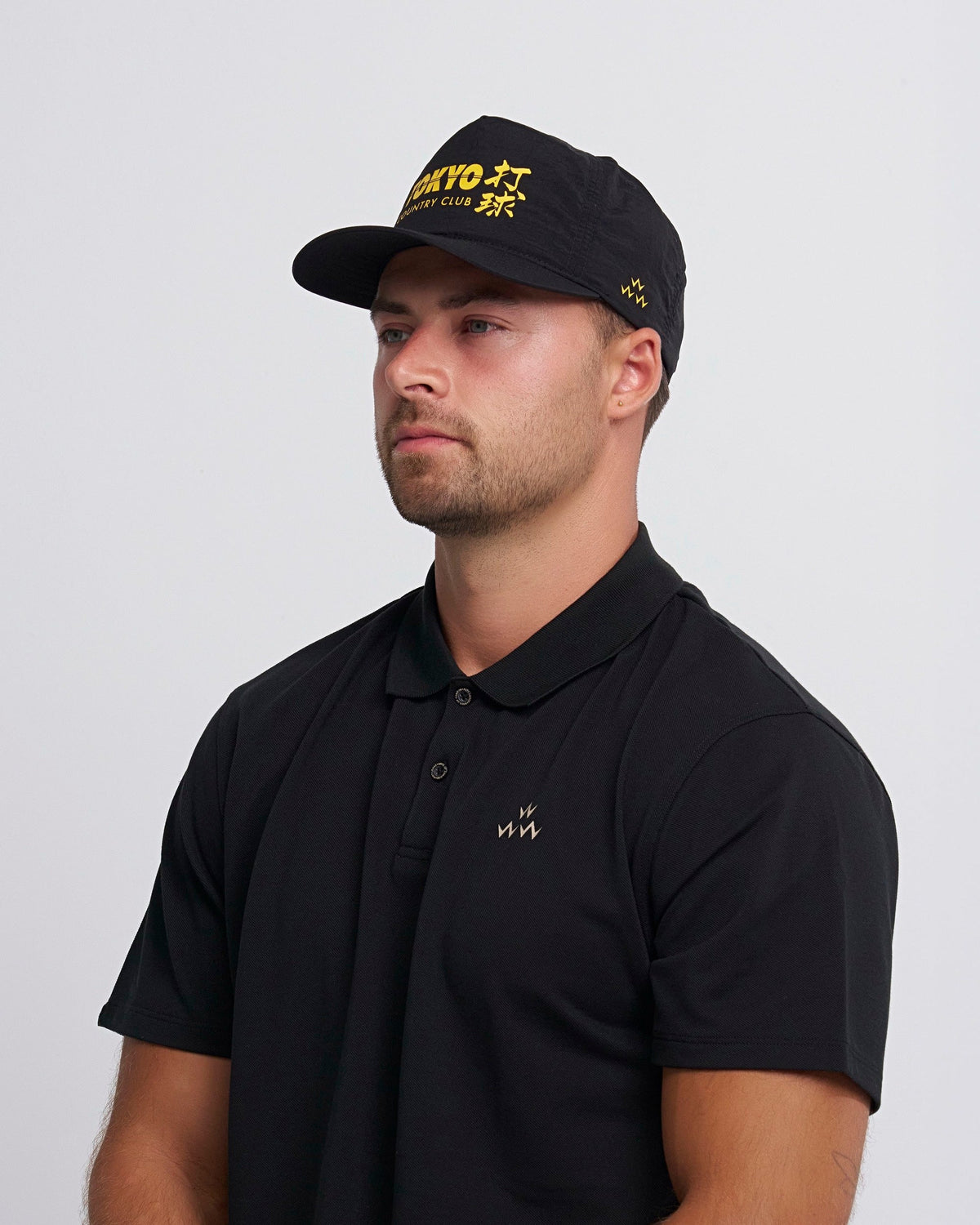 A man wearing a black 5-panel cap with yellow Japanese text and a matching black polo shirt.