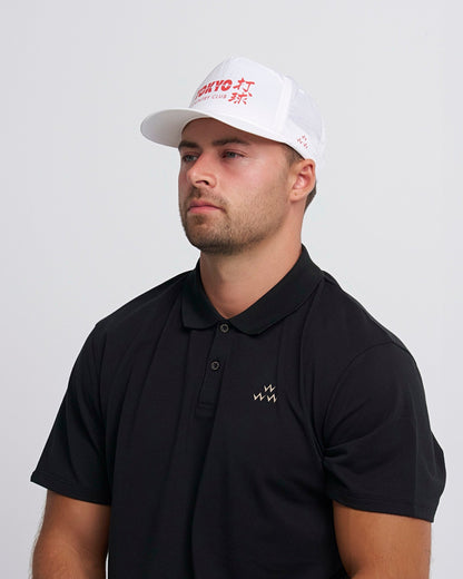 A man wearing a white snapback with a red Tokyo country club logo print, and a black polo shirt.