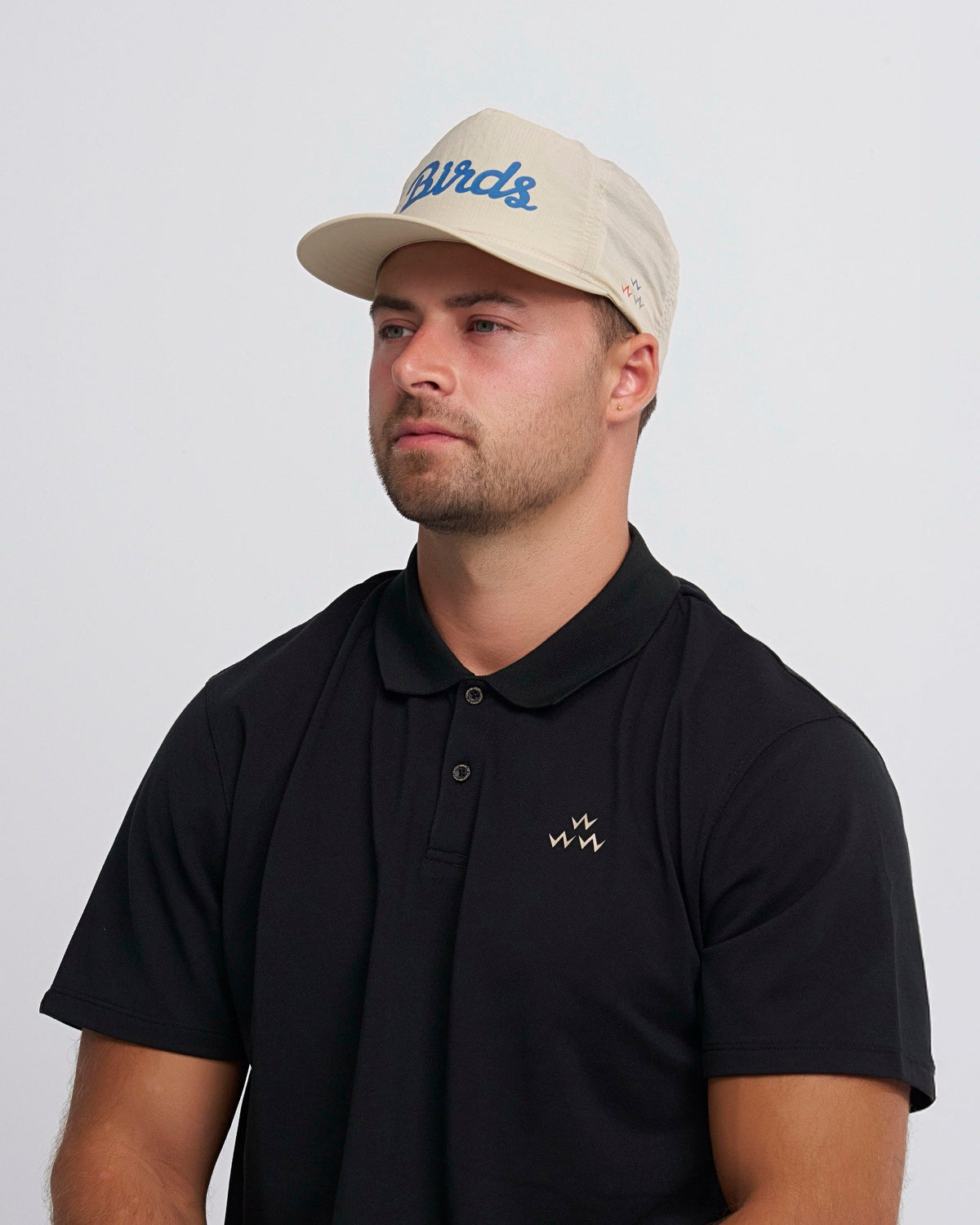 A man wearing a tan snapback cap with a blue script design and a black polo shirt with a small chest logo.