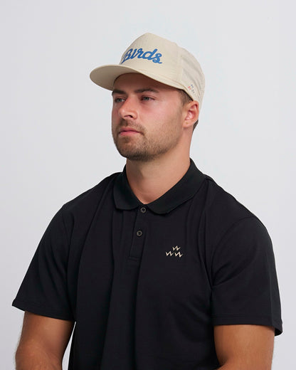 A man wearing a tan snapback cap with a blue script design and a black polo shirt with a small chest logo.