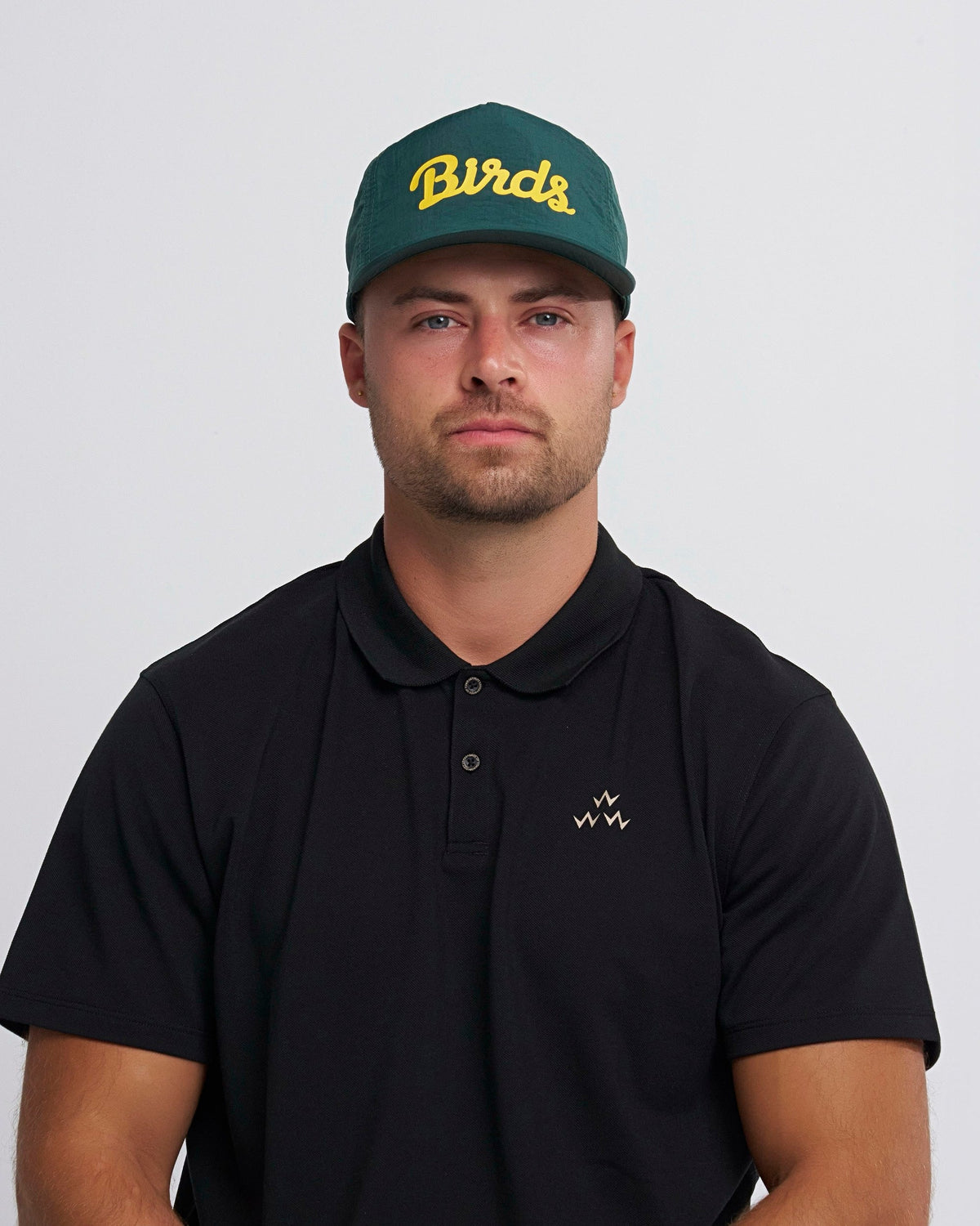 A man wearing an alpine green nylon snapback cap with a yellow Birds script print and curved brim.