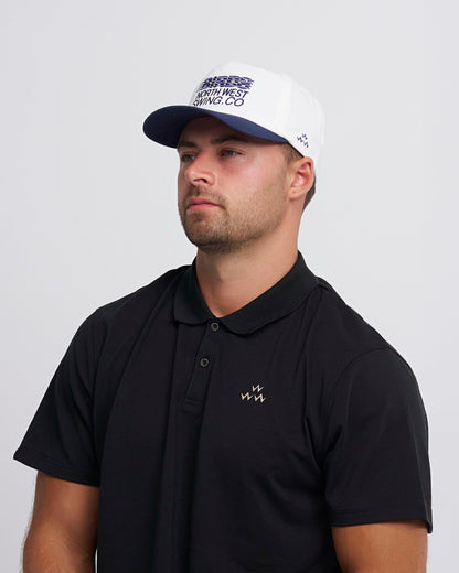 A man wearing a white snapback cap with a navy brim and blue text embroidery on the front.