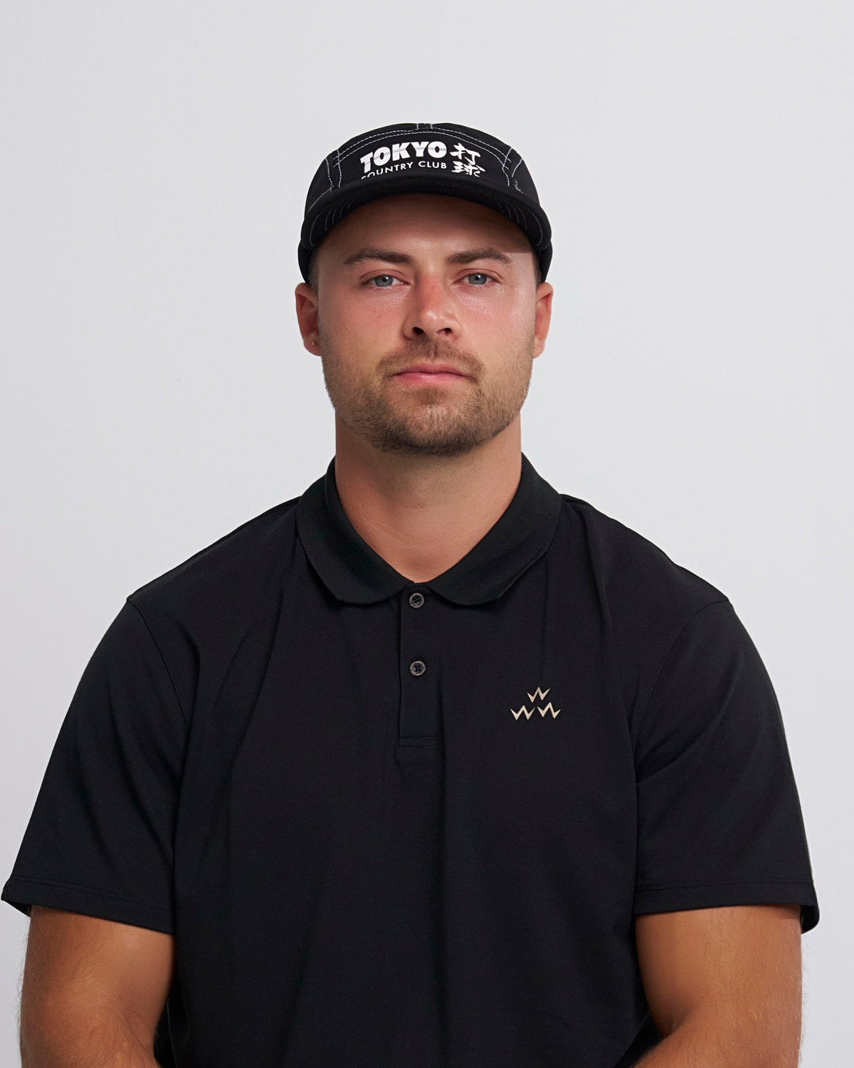 Man wearing a black polo shirt with a logo and a cap with 'Tokyo Country Club' text on a light gray background