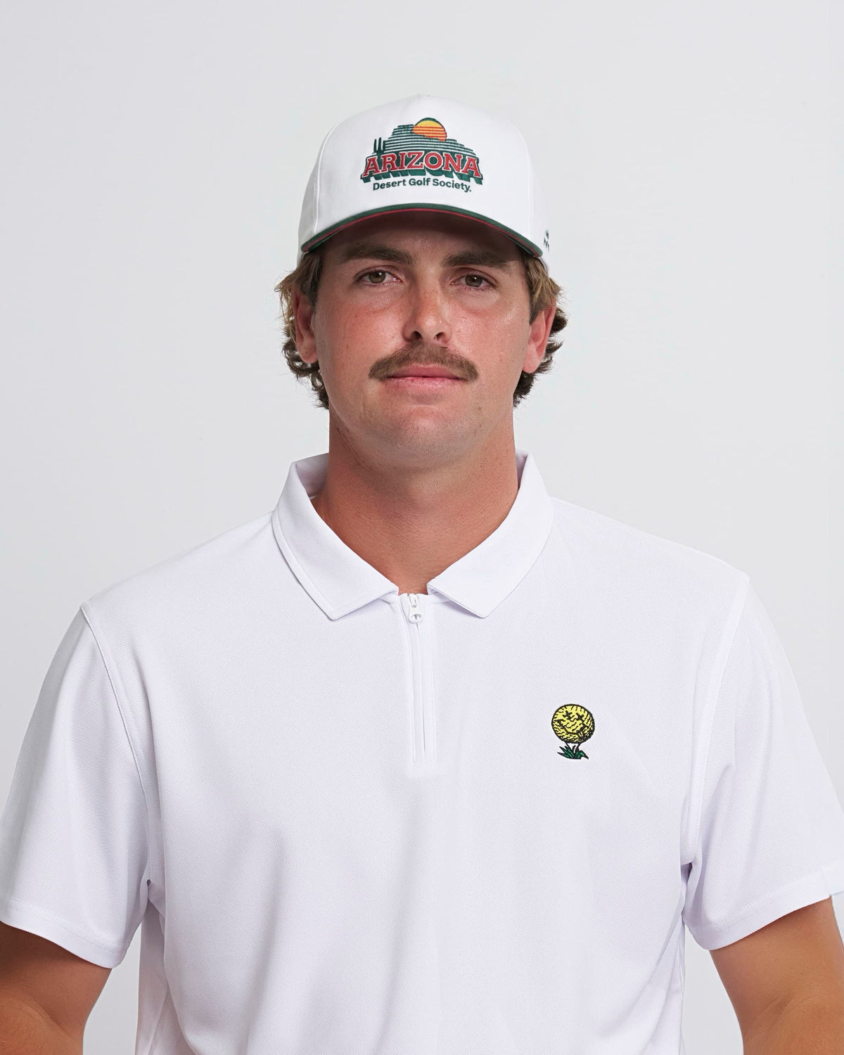 A man wearing a white snapback with a green brim and a sunset desert landscape graphic on the front.