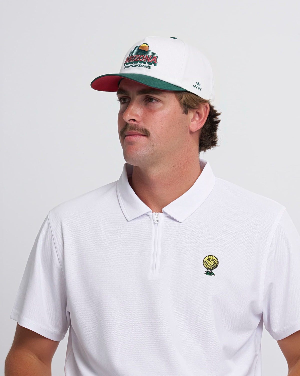 A man wearing a white snapback hat with a green curved brim and a colorful Arizona Desert Golf Society graphic.
