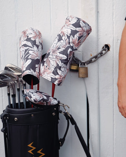 A black golf bag holding clubs fitted with azalea print headcovers, next to a person's arm.