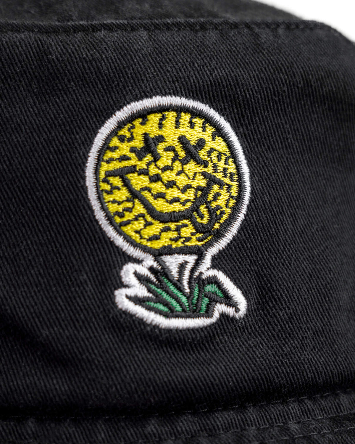 Detail of a black cotton boonie featuring a smiley face golf ball design atop green grass.