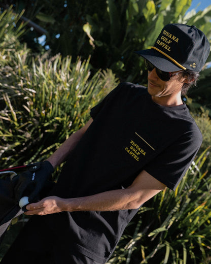 A man wearing a black t-shirt with embroidered yellow chest text, and a matching black cap outdoors.