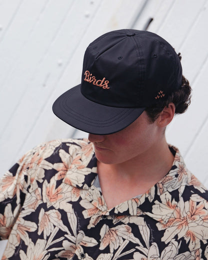 A man wearing a black snapback hat with embroidered script lettering and side bird icon.