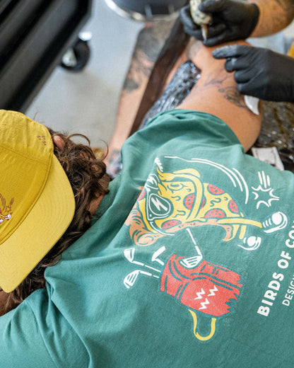 A person wearing a gold snapback cap while receiving a tattoo.