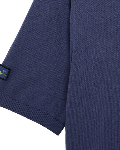 Detail of a crown blue cotton knit polo sleeve with a ribbed cuff and a small woven logo patch.