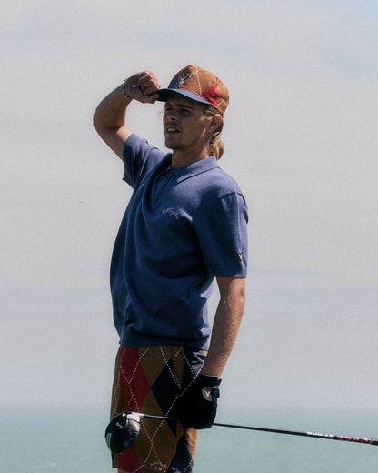 A man wearing a crown blue polo shirt, patterned shorts, and a cap while holding a golf club outdoors.