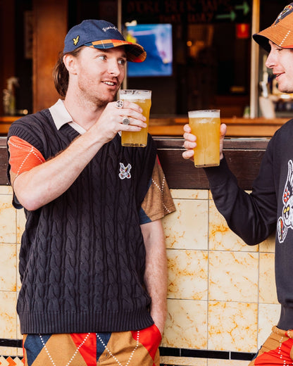 Two men drinking beer, one wearing a black cable knit vest with an embroidered winged golf ball.
