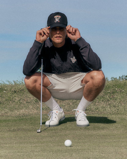 A man wearing a black cable knit vest, matching cap, and beige shorts, crouching on a green golf course.