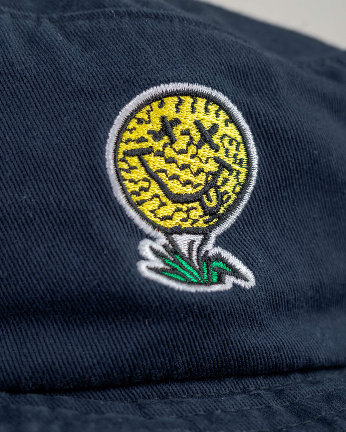 Detail of a navy cotton boonie featuring a yellow smiley face golf ball logo with X eyes on a tee.