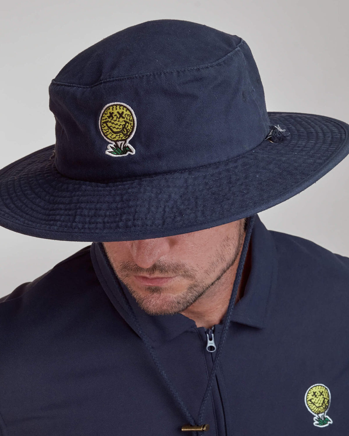 A man wearing a navy boonie hat with a yellow smiley golf ball graphic and a tonal drawcord.