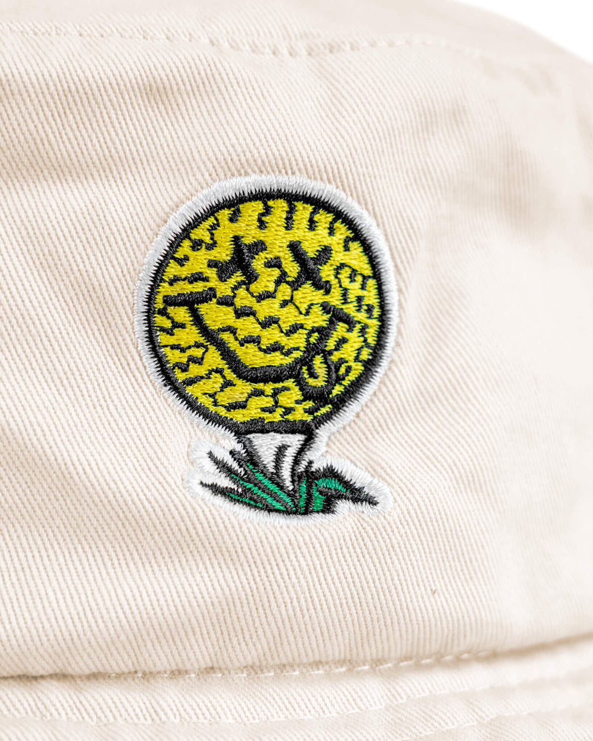 Detail of off-white cotton canvas boonie featuring a smiley face golf ball logo on a tee in grass.