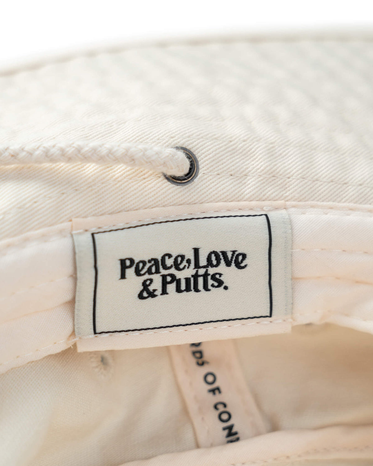 Detail of an off-white boonie with a woven label inside reading Peace, Love & Putts and a drawcord.