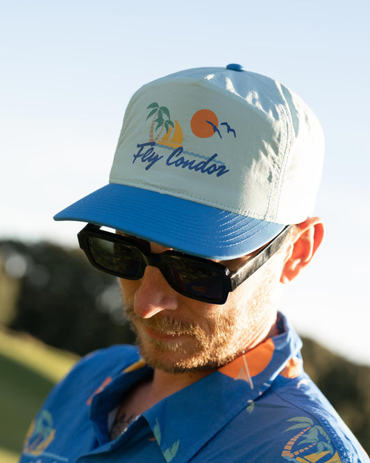 A man wearing a sky blue five-panel nylon snapback cap with a sunset beach graphic and Fly Condor text.