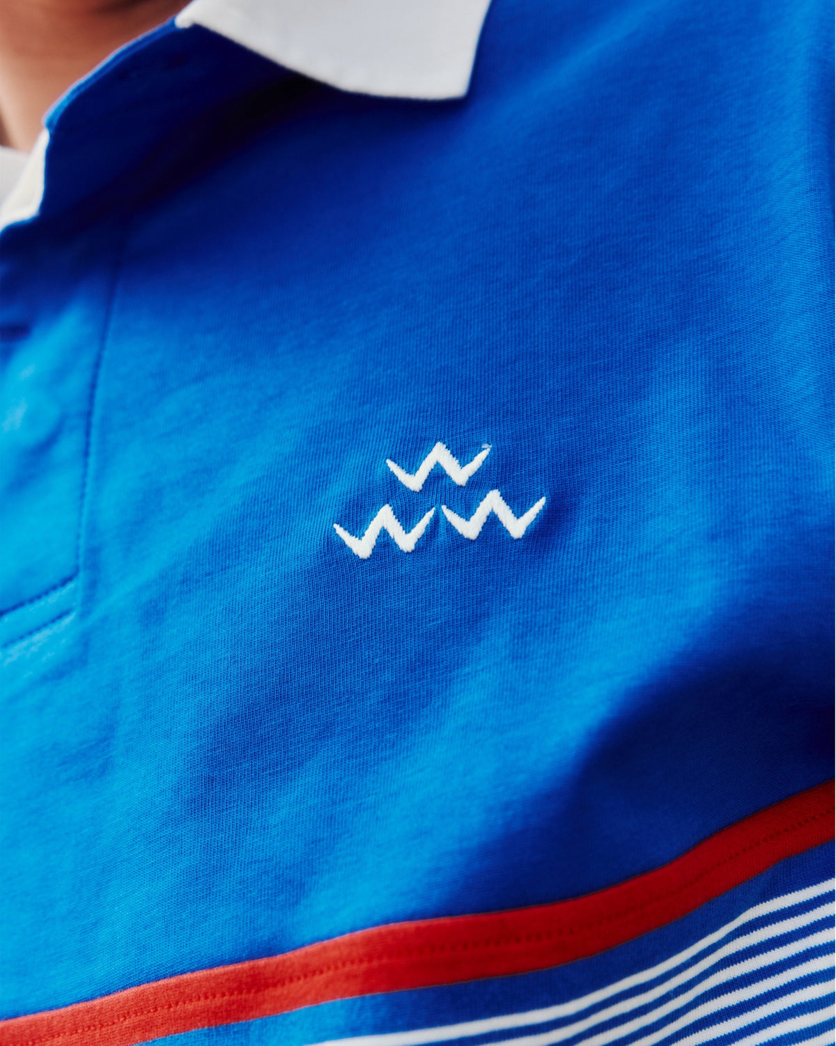 Detail of a blue sweater with a white collar, embroidered white bird logo, and red stripe.