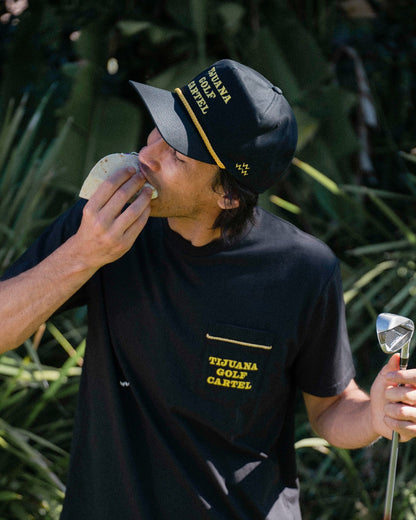 A man wearing a black snapback with yellow lettering and a rope across the brim while eating a taco.