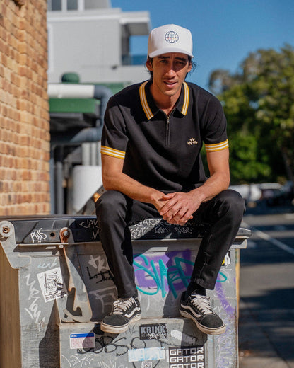 A man wearing a black knit polo shirt with a yellow striped collar, quarter zip, and a triple eagle logo.