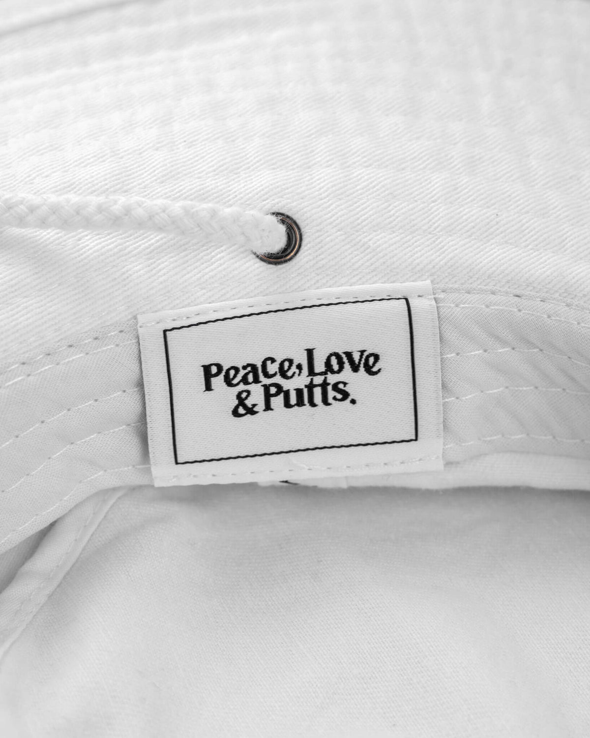 Detail of a white cotton canvas boonie featuring a tonal drawcord, brass fastener, and a label reading Peace, Love & Putts.