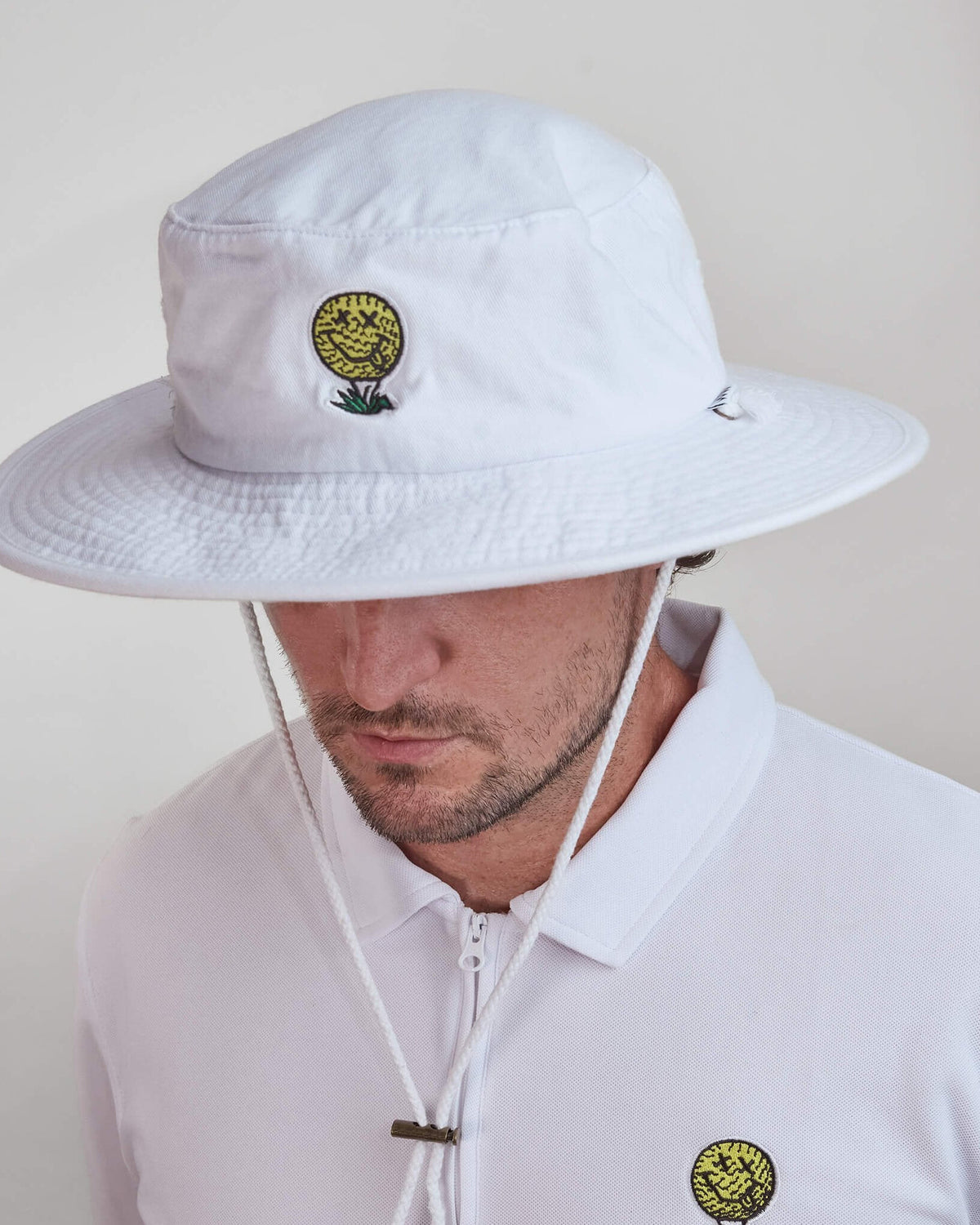 A man wearing a white cotton boonie hat featuring a yellow smiley face golf ball design on the front.