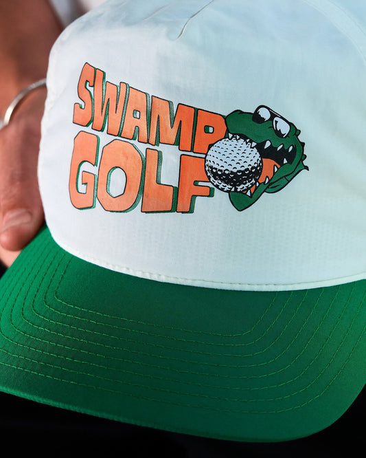 A person holding a white nylon cap featuring an orange Swamp Golf text and a green alligator design.