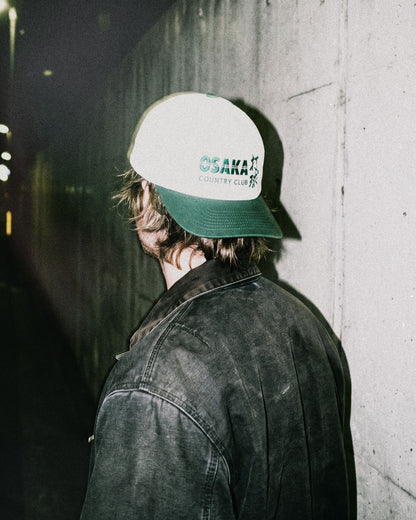 A man wearing a white snapback hat with a green brim and green Osaka Country Club text on the front.