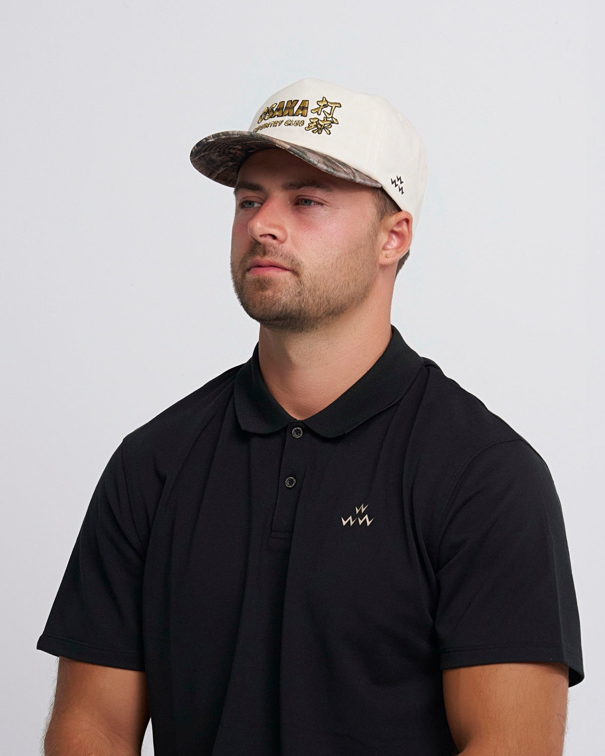 A man wearing a natural snapback cap with a camo brim and gold lettering on the front.