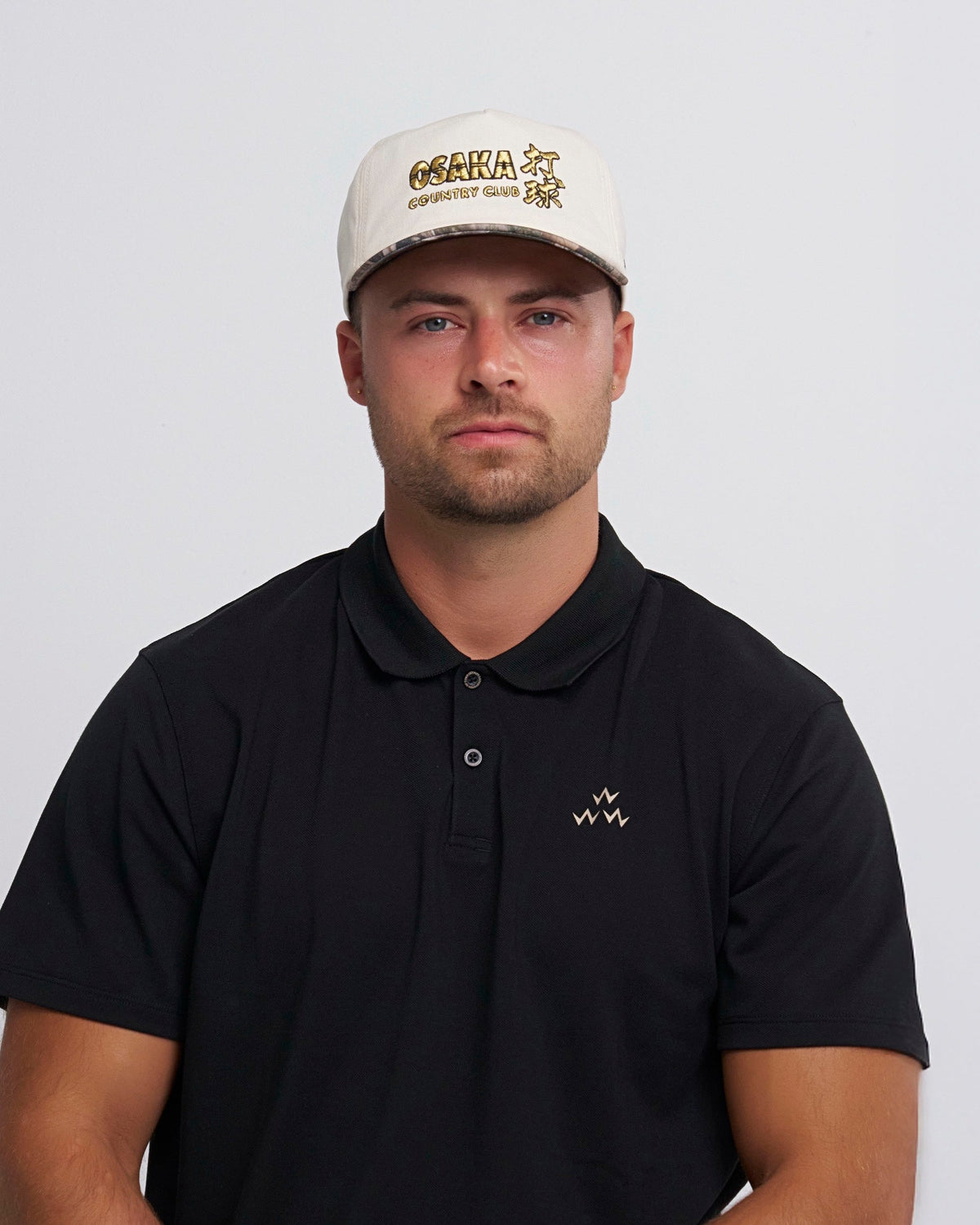 A man wearing a natural snapback cap with a gold embroidered graphic and a camo brim.