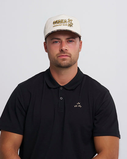 A man wearing a natural snapback cap with a gold embroidered graphic and a camo brim.
