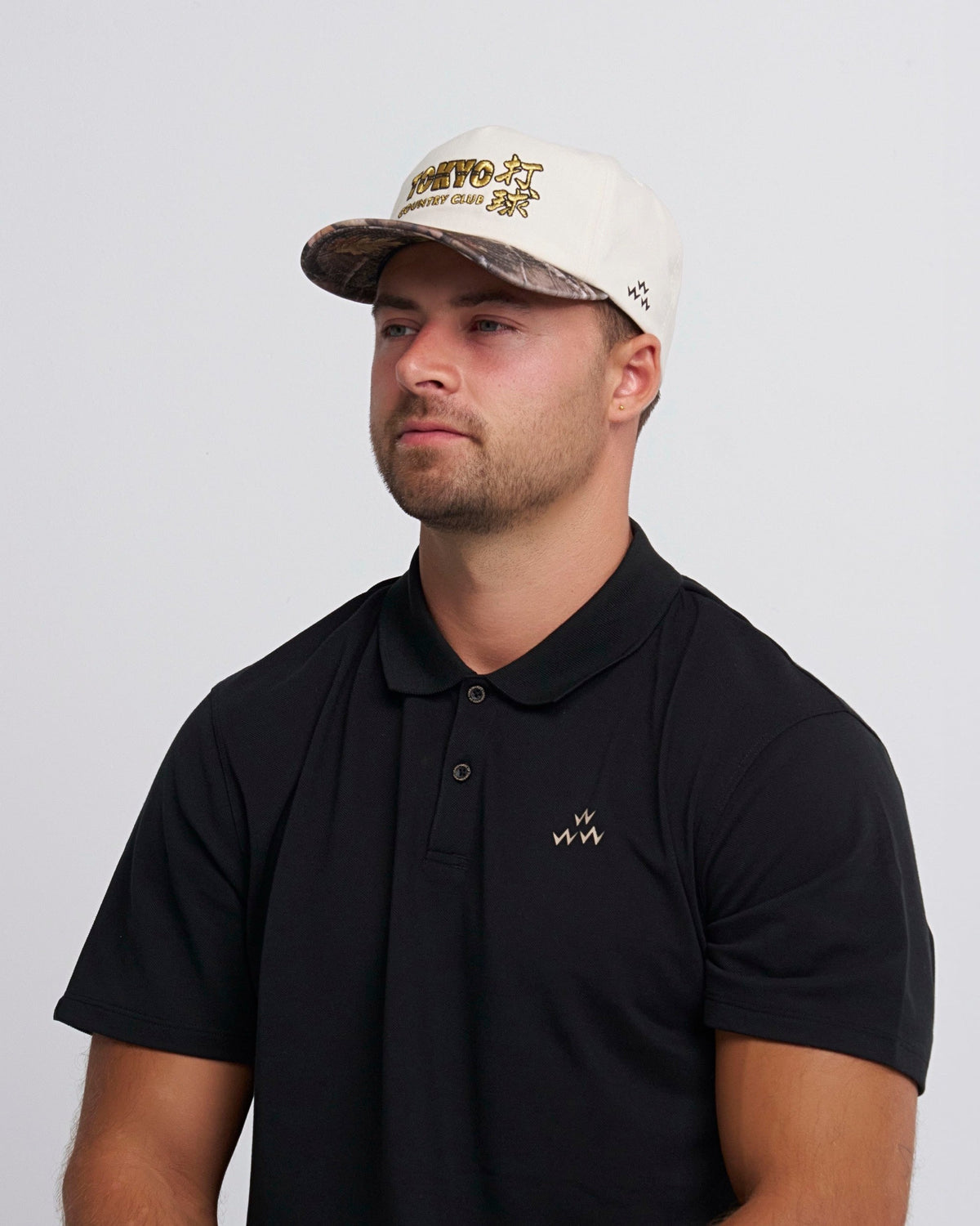 A man wearing a natural snapback cap with a camo brim and gold Tokyo Country Club embroidery.