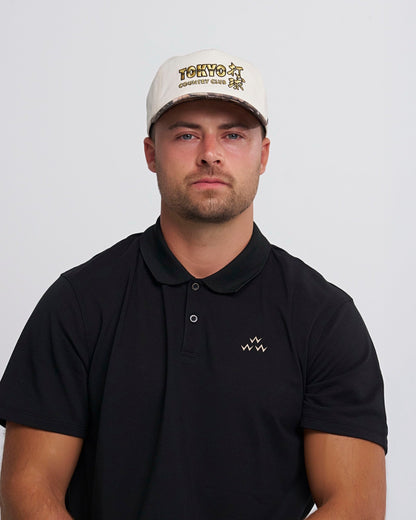 A man wearing a natural snapback cap with a gold Tokyo Country Club embroidered graphic on the front.