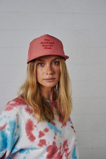 A woman wearing a coral snapback cap with The Weekend Rescue Club printed in red on the front.