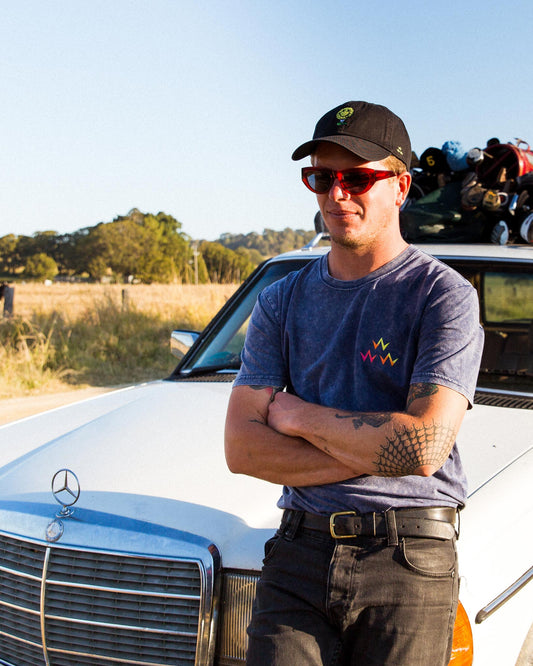 A man wearing a black cap with a golf ball motif, red sunglasses, and a grey tee, leaning on a car.