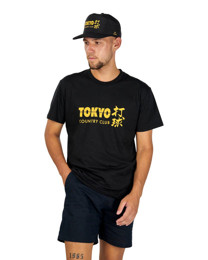 A man wearing a black t-shirt with a yellow Tokyo Country Club graphic, black shorts, and a matching cap.