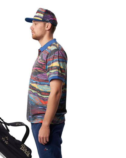 A man wearing a multi-coloured Coogi print cap, matching polo shirt, and navy blue trousers.