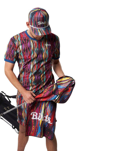 A man wearing a colourful Coogi print t-shirt, shorts, and a cap while holding a matching golf towel.
