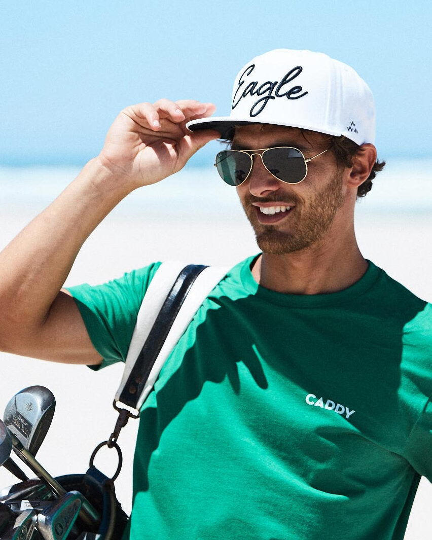 A man wearing a white snapback hat with black cursive text and a flat black undervisor on a sunny beach.